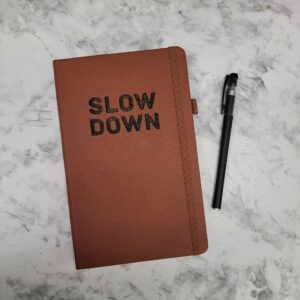 Brown leather journal with slow down burned into the cover and a black pen sitting on a marble background